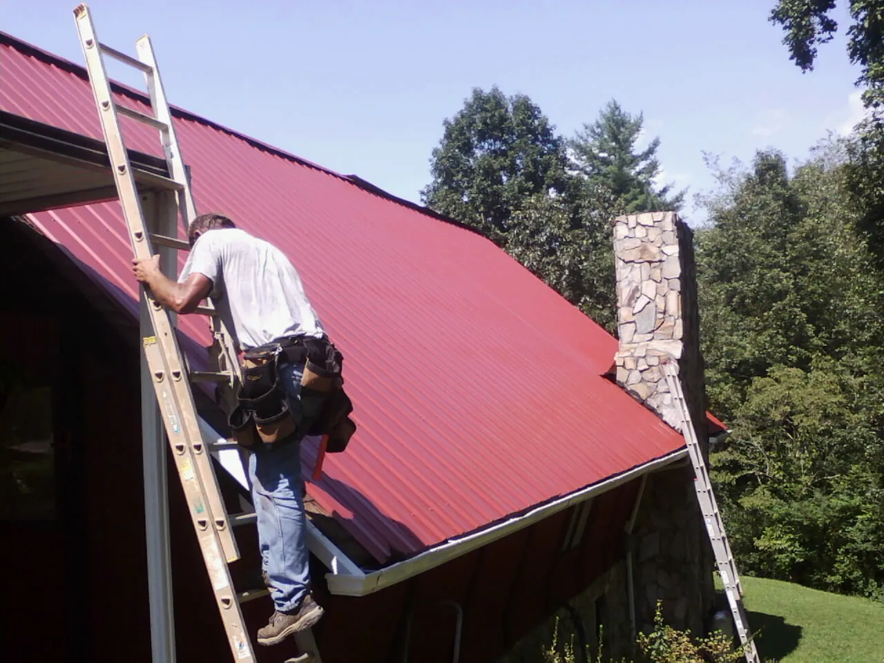 Roofer on a ladder beside a red metal roof and stone chimney — Soffit & Fascia Repair by Roofing Brothers in Duluth